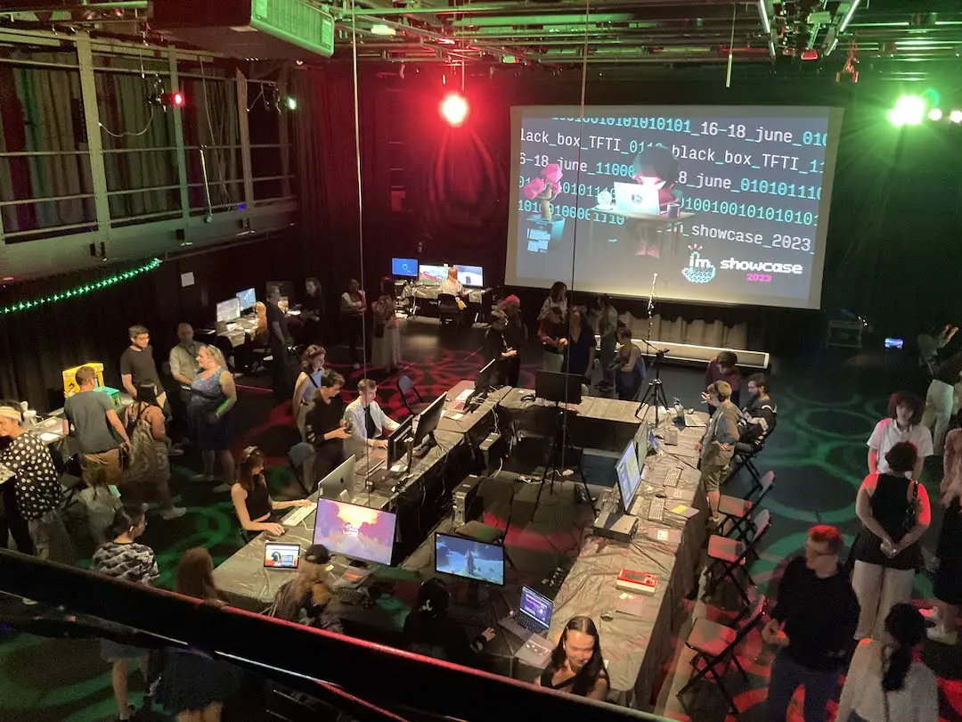 A birdseye view of the event while in full swing. People mill around computers at the edge of the room and on a central island. At the far end an animation of a duck hacking in front of some binary code is projected onto a canvas.